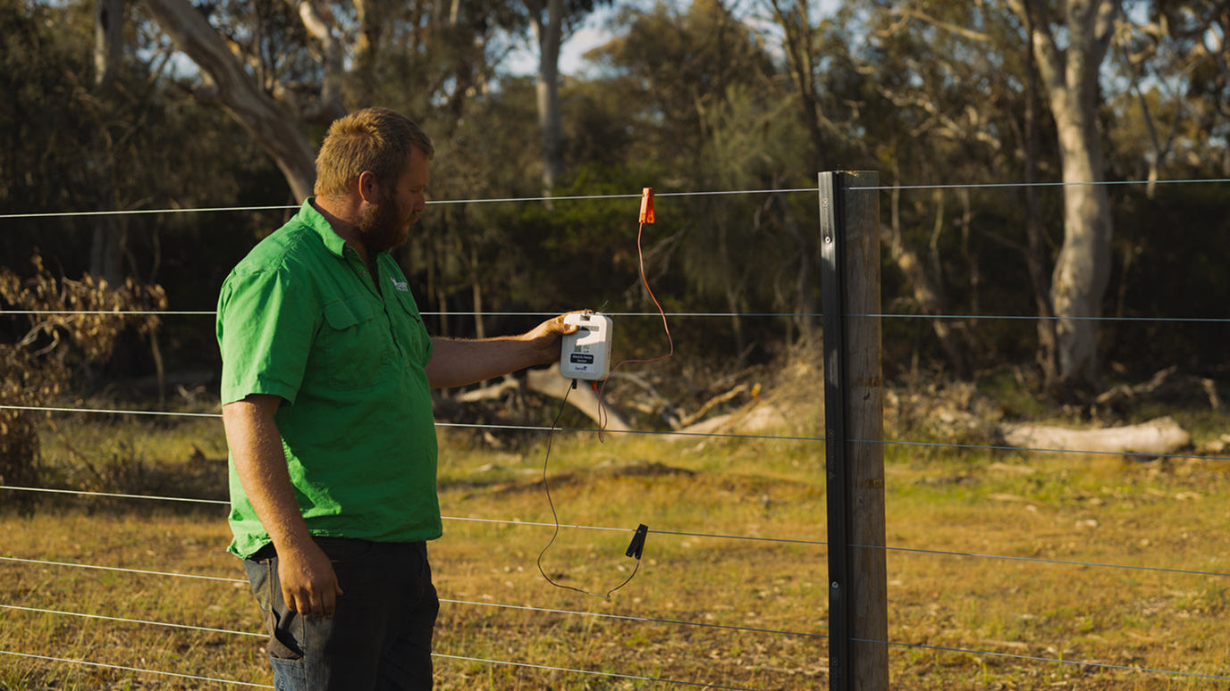 Man in a green shirt adjusting electric fence sensor in a rural setting