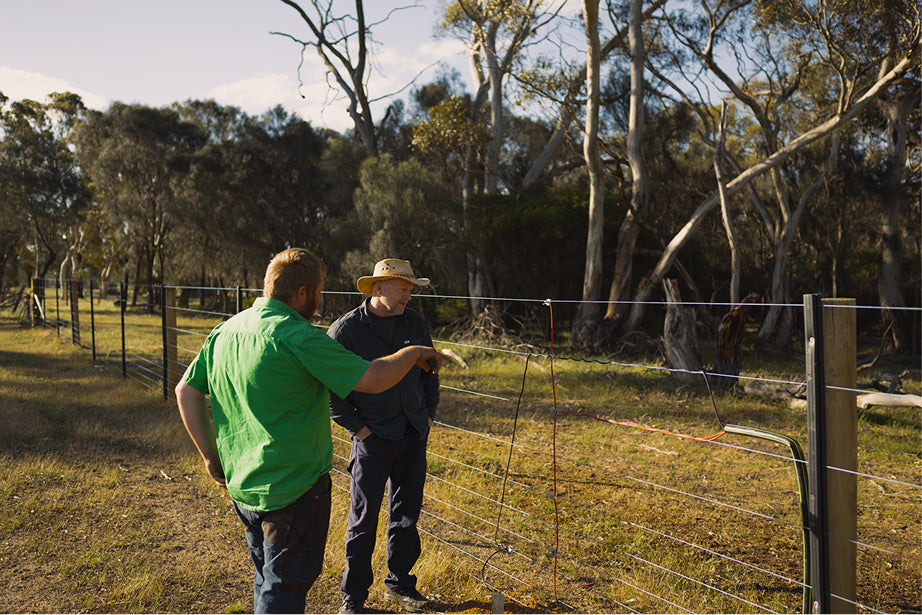Two men standing near an electric fence in a rural setting with trees in the background.